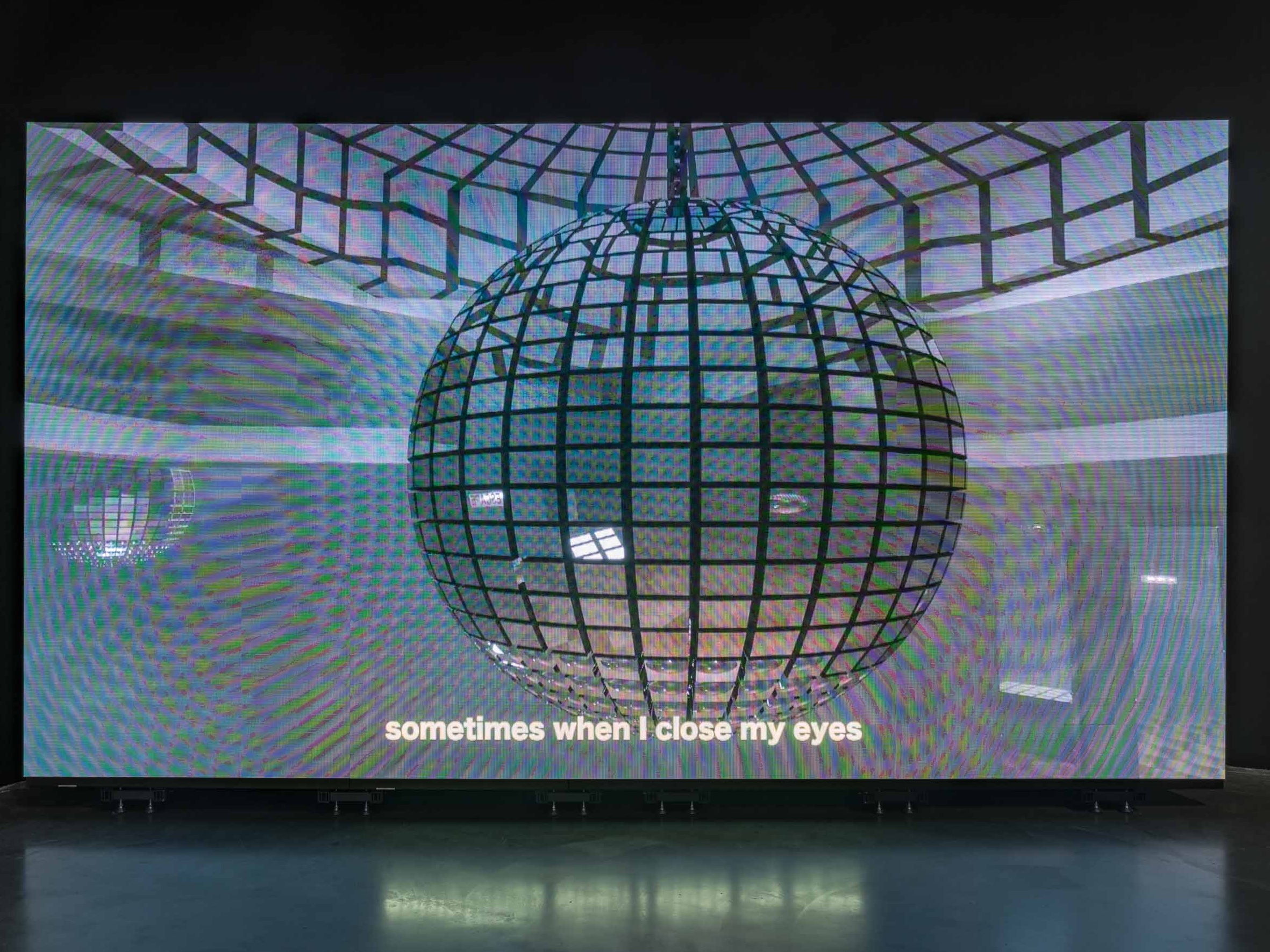 Image of a large projection screen glowing in the dark. The Projection screen plays an animated, 3-D rendering of a disco ball. The bottom of the screen has a subtitle that says "sometimes when I close my eyes."